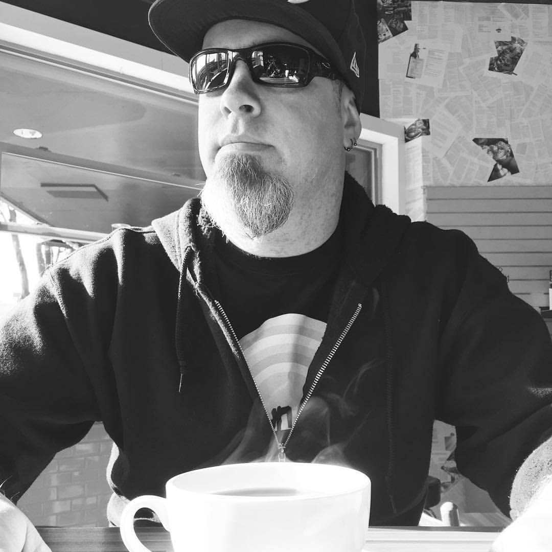 A black and white picture of Jim sitting at a table, wearing a sweatshirt, baseball hat, and sunglasses. Jim looks to the left and out of frame over the top of a steaming cup of coffee.