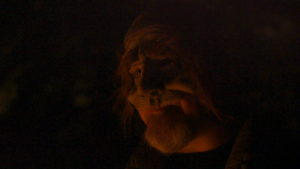 A dark picture of a homeless man in an old man mask. This is a close-up shot of the face of the actor only, and he seems to be lit with a red light.